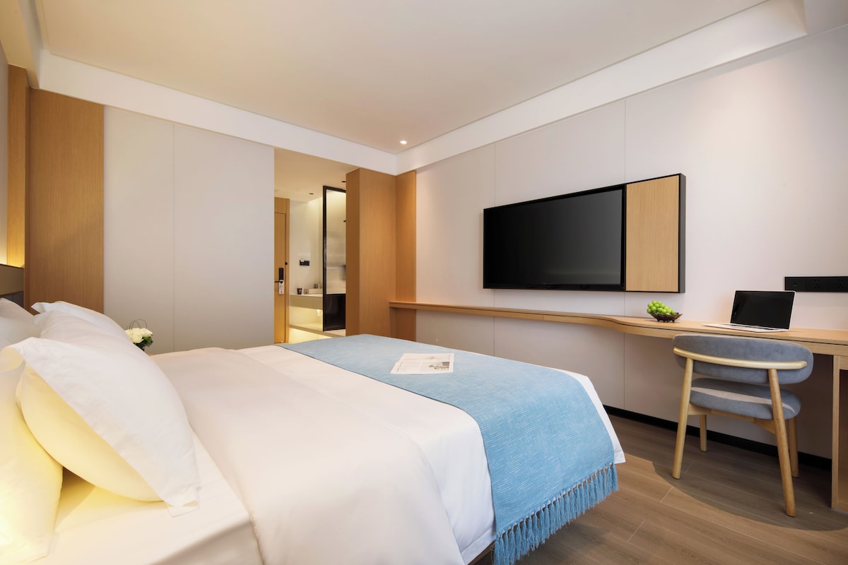 A hotel room with a bed, a desk, a chair, and a flat-screen TV mounted on the wall.
