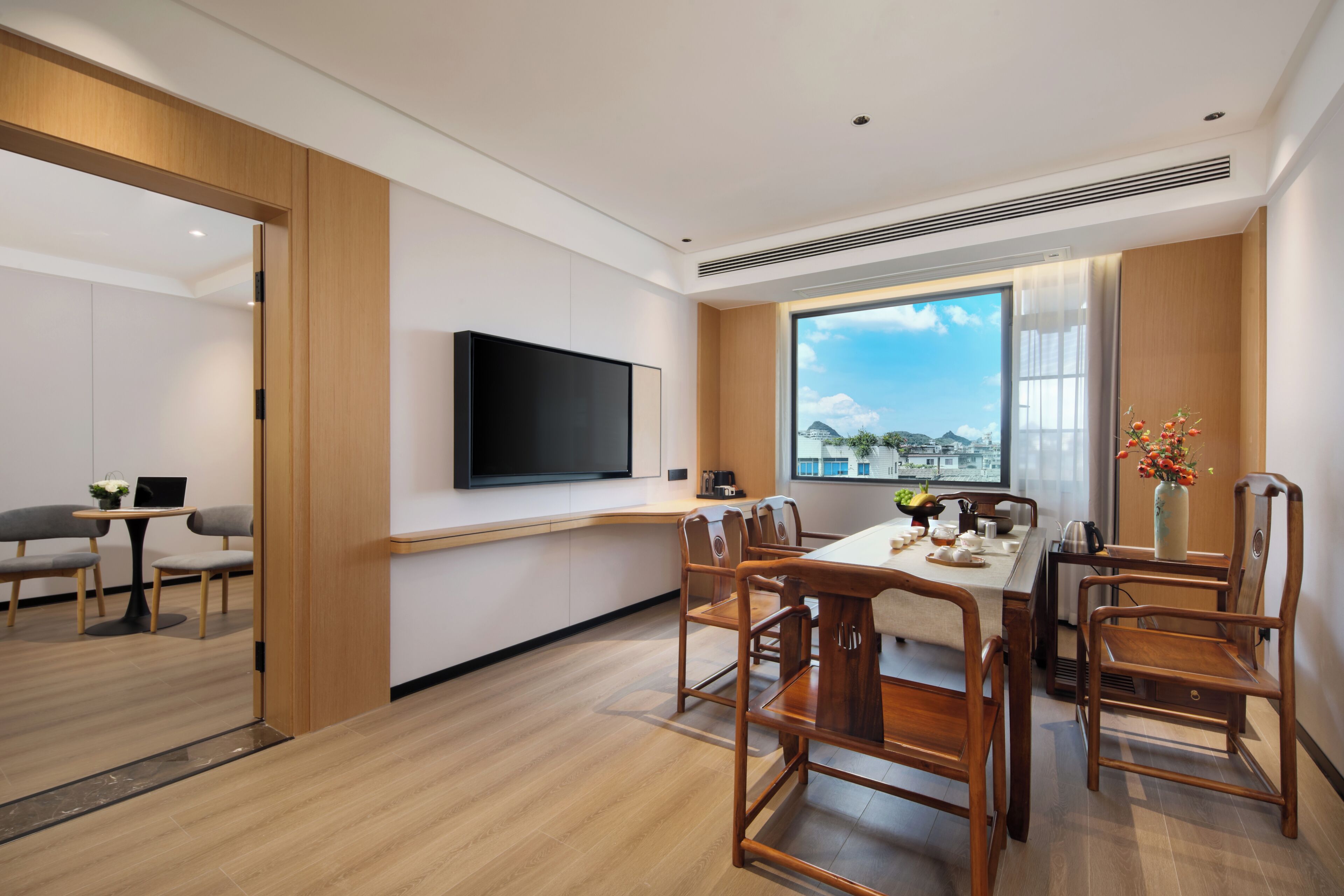 A modern hotel room with a dining area, a flat-screen TV mounted on the wall, and a large window offering a view of the cityscape.
