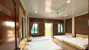 Habitación de hotel con tres camas individuales, ventilador de techo y vistas al exterior a través de una ventana.