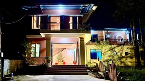 Una casa de dos pisos con un balcón, una escalera que lleva a la entrada y un patio delantero bien iluminado.