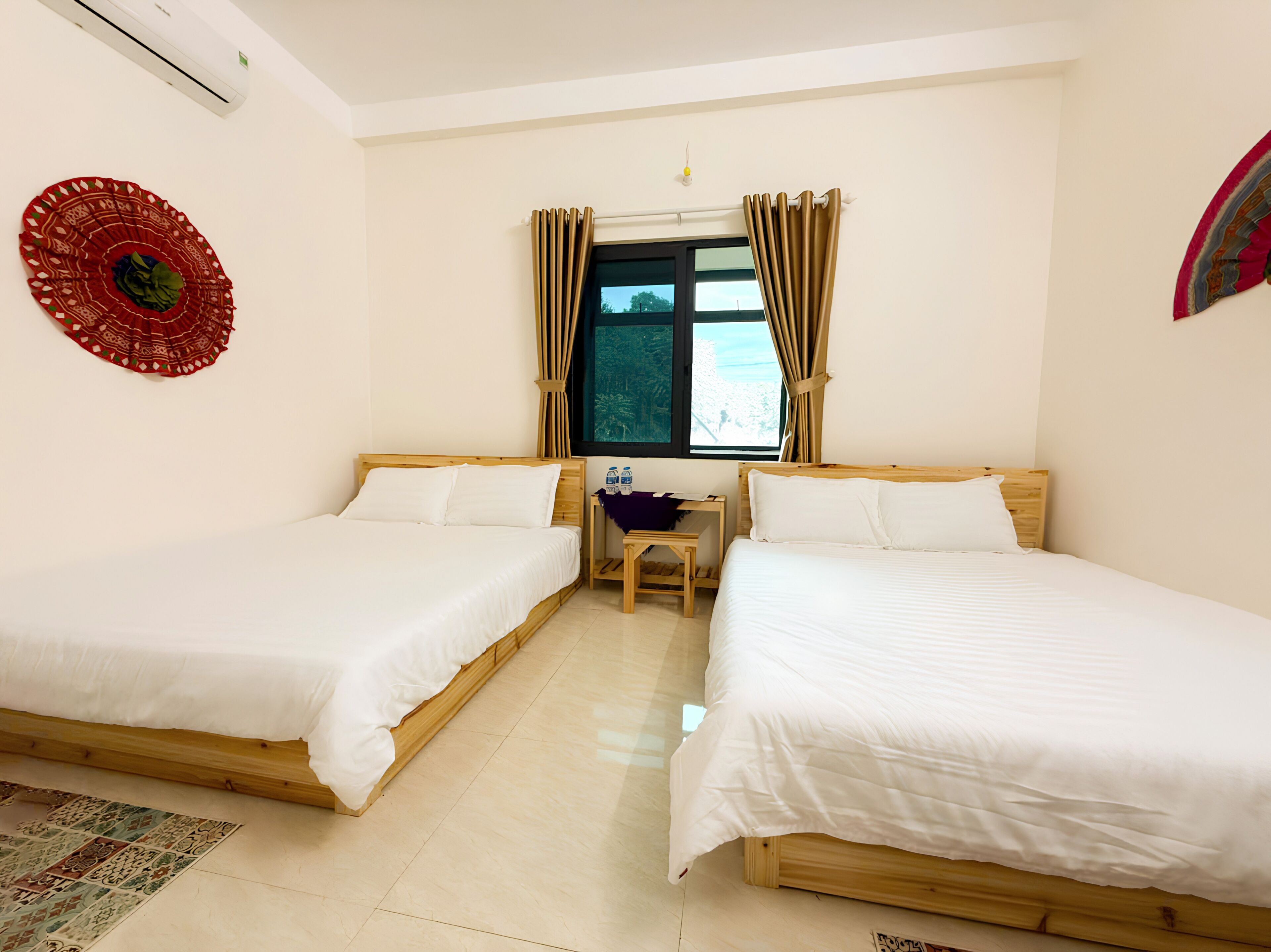 Una habitación de hotel con dos camas individuales, una mesita, una silla, una ventana con cortinas y un aire acondicionado instalado en la pared.