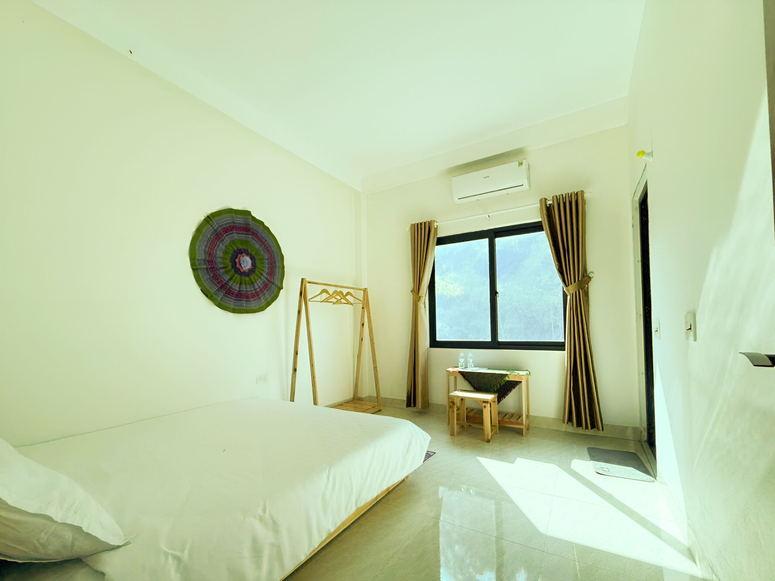 Une chambre d’hôtel comprenant un lit, une petite table, une fenêtre avec des rideaux, un appareil de climatisation et une assiette décorative fixée au mur.