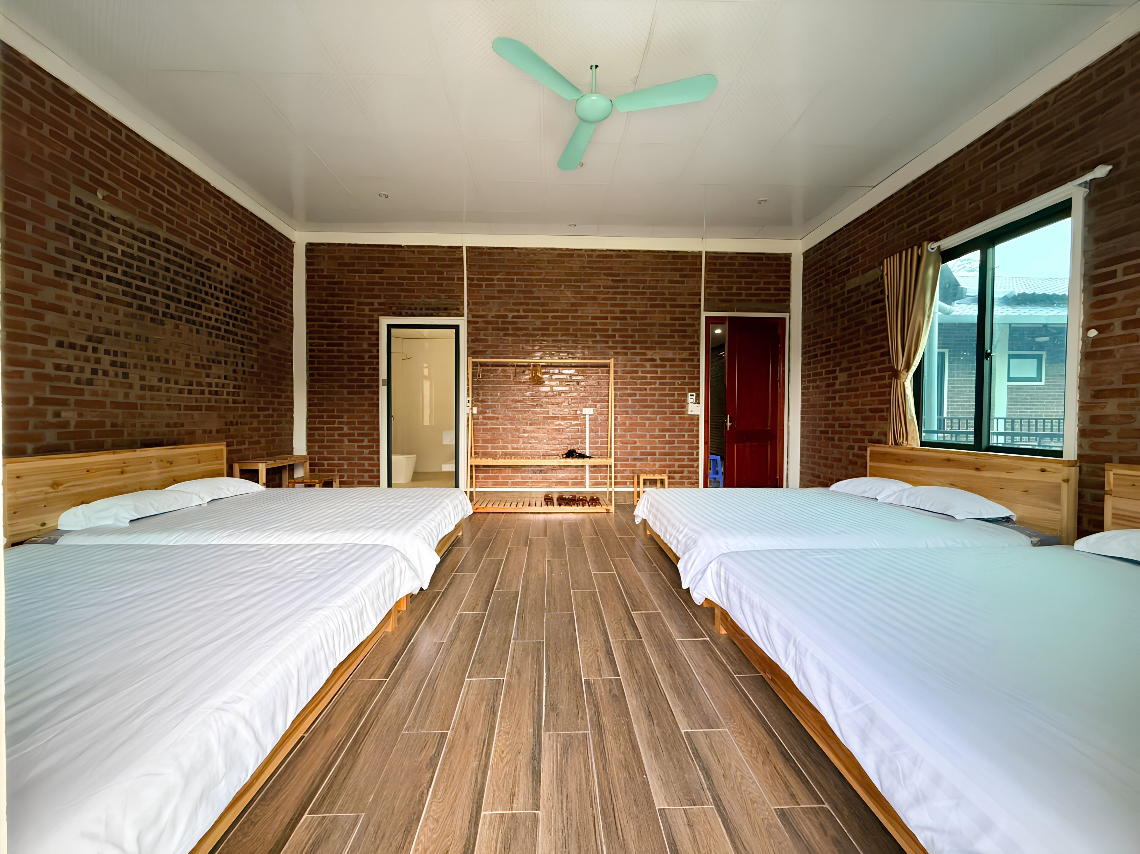 Une chambre d’hôtel avec deux lits, un plancher en bois, des murs en briques, un ventilateur au plafond et une fenêtre avec des rideaux.