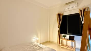 A bedroom with a bed, an air conditioning unit, a small table with a plant, and a window with curtains.