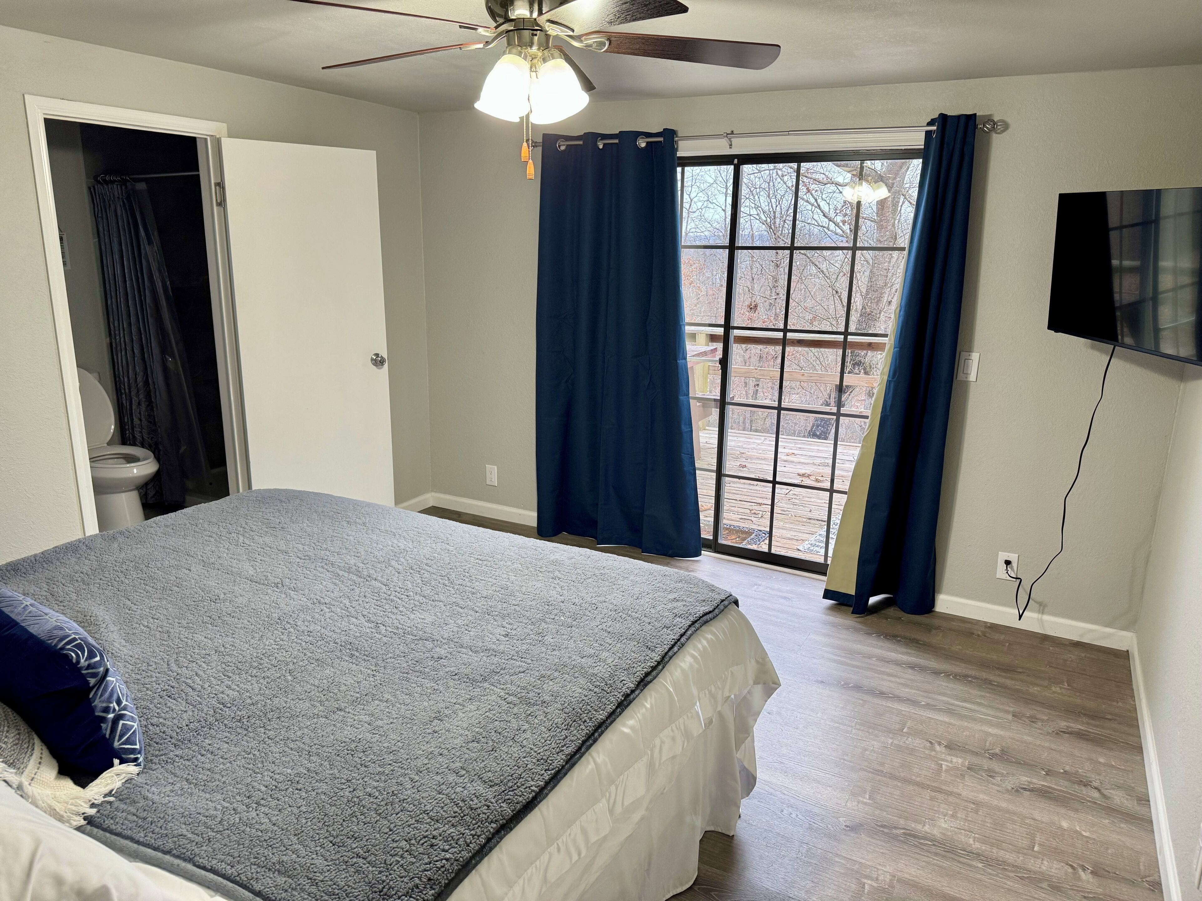 A bedroom with a bed, a ceiling fan, a window with curtains, and a TV mounted on the wall.