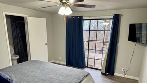 Une chambre à coucher avec un lit, un ventilateur de plafond, une fenêtre avec des rideaux et un téléviseur fixé au mur.