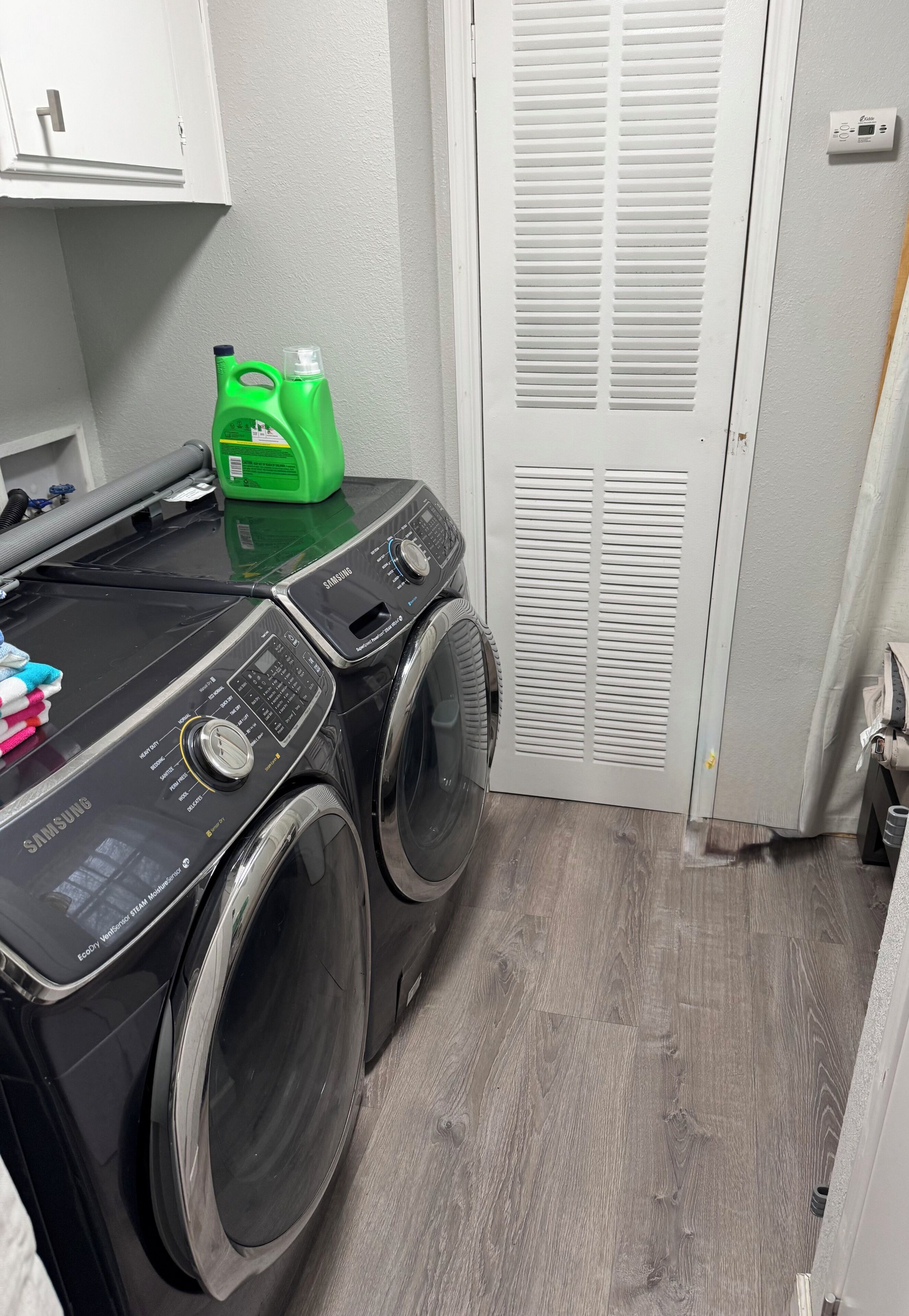 A laundry area with a Samsung washing machine, a green detergent bottle, and a white door with a vent.