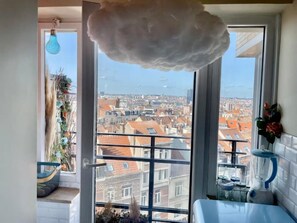 A room with a large window, a hanging light fixture, a balcony with a railing, and a kitchen area with a blender.
