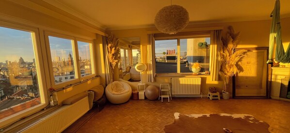 A room with a large window, a radiator, a round chair, a small stool, and a hanging light fixture.