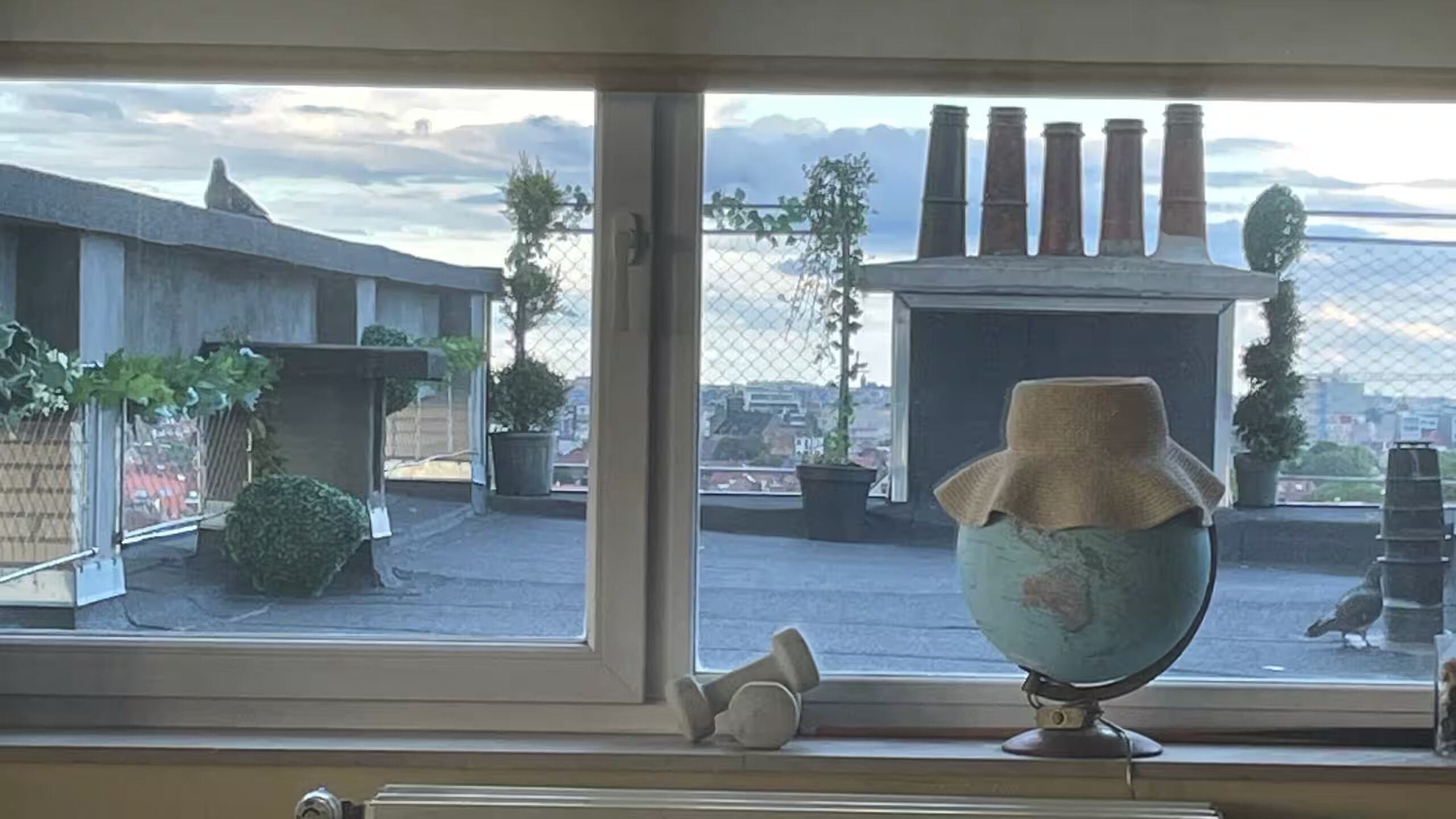 Une vue à travers une fenêtre, avec un globe décoratif, une sculpture en métal et un espace extérieur clôturé agrémenté de plantes en pot.
