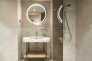 Un baño moderno con ducha envuelta en vidrio, un espejo redondo con luces LED y un mueble de tocador de madera.