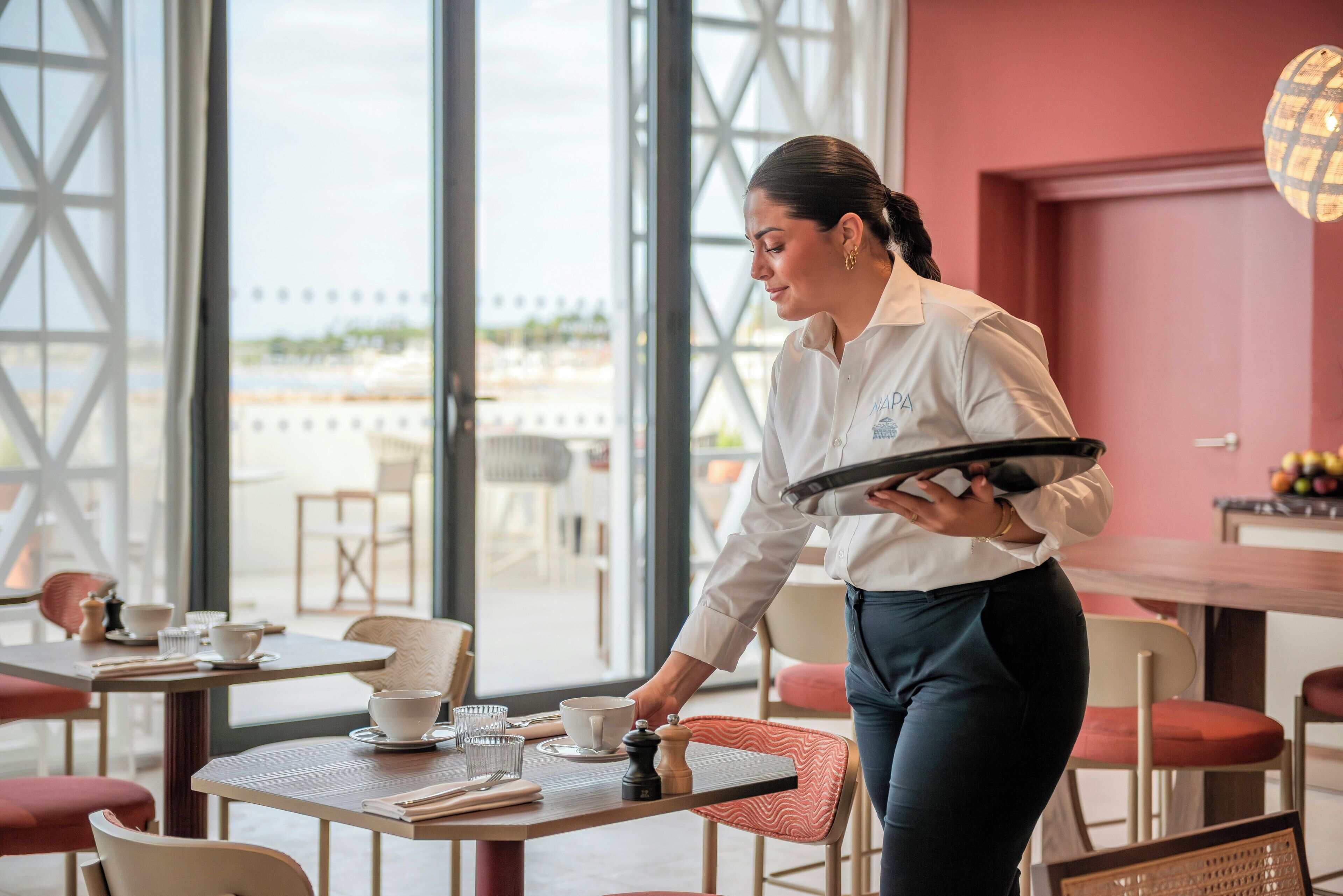 Eine Person bedient ein Tablett mit Gläsern und einer Tasse in einem Raum mit großen Fenstern und Aussicht.