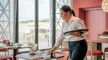 Eine Person bedient ein Tablett mit Gläsern und einer Tasse in einem Raum mit großen Fenstern und Aussicht.