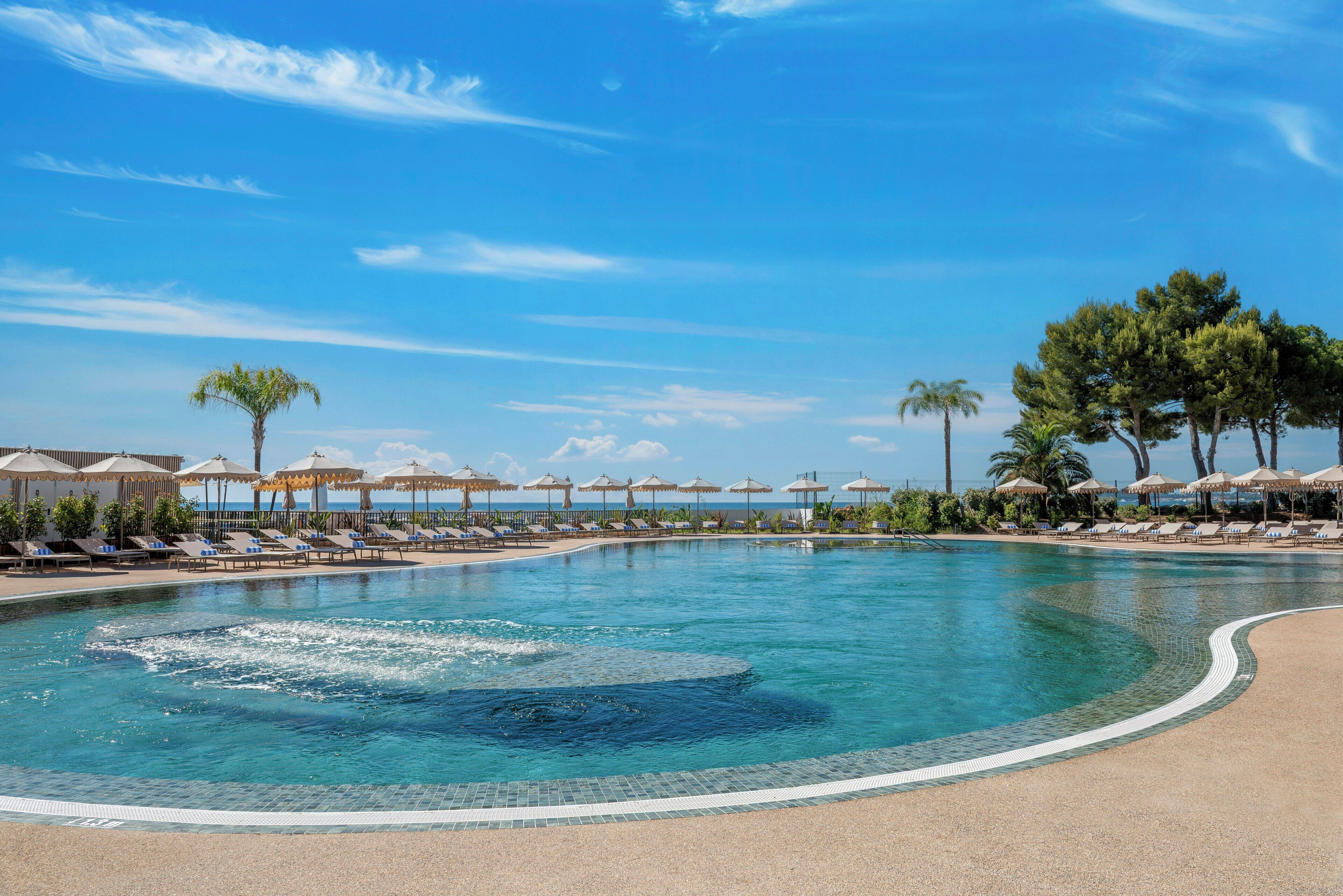 Un área de piscina con sillones de descanso y sombrillas, rodeada de árboles y un cielo despejado.