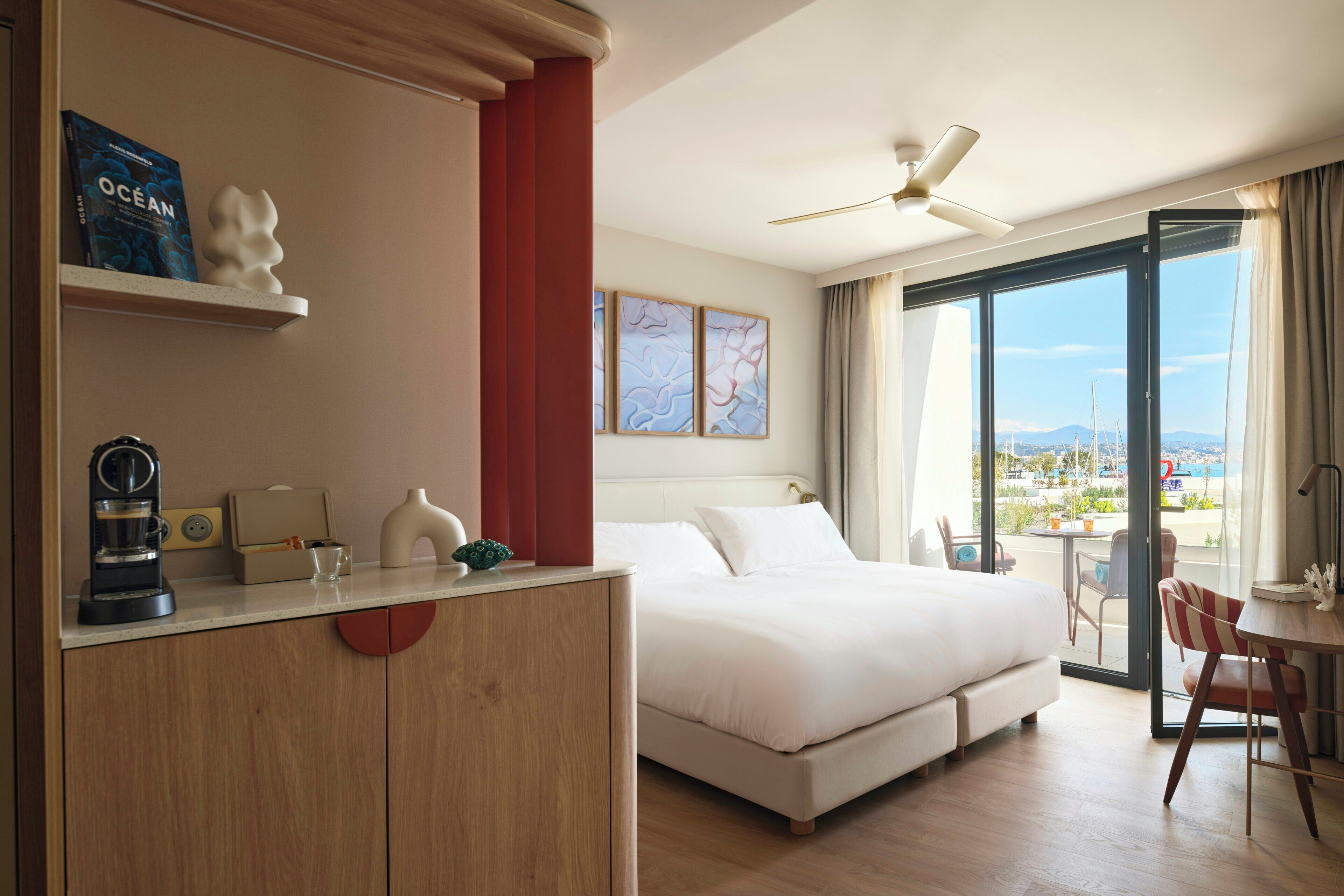 A hotel room with a bed, a desk, a coffee maker, a view of the sea, and a balcony with chairs.