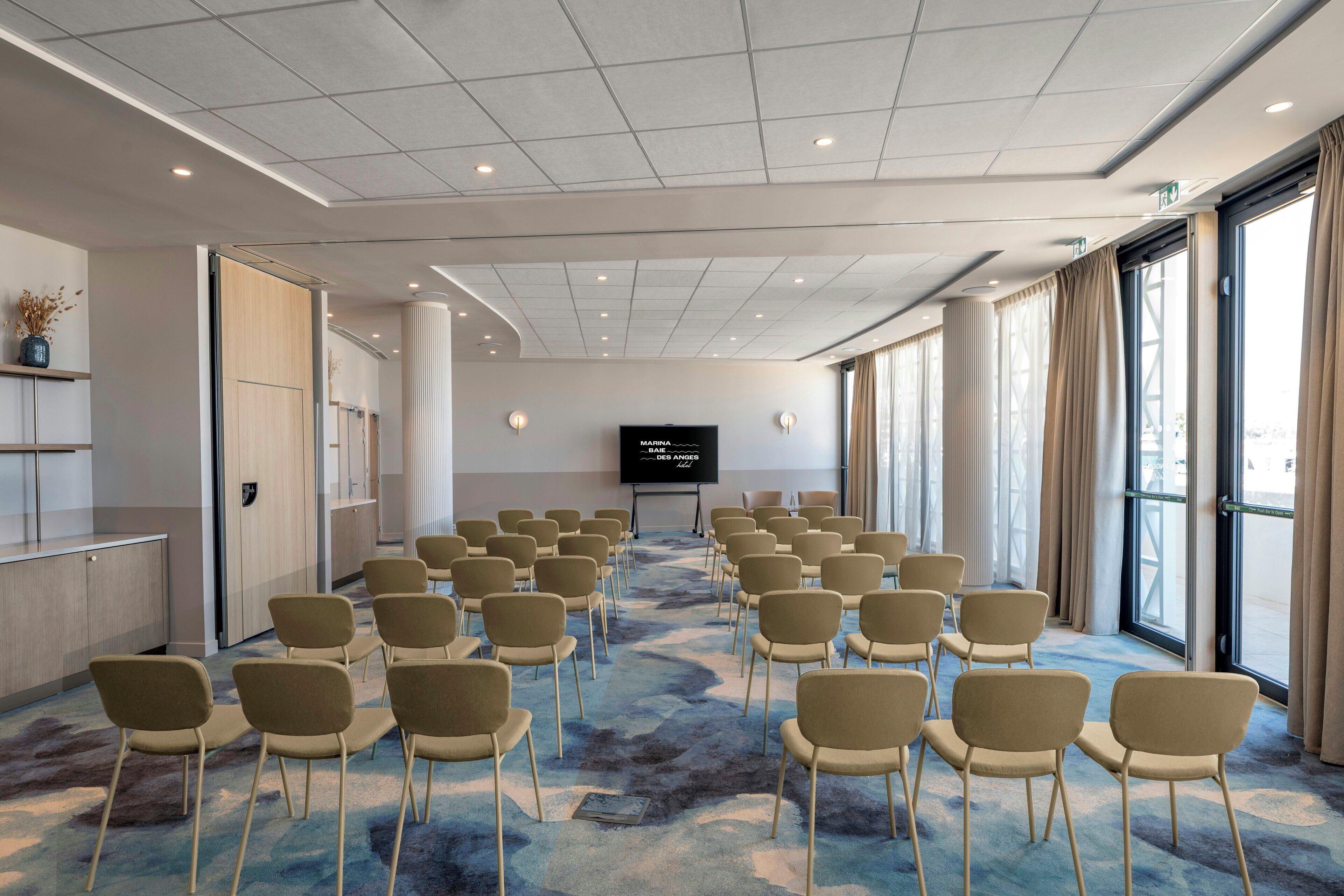 Uma sala de conferências com fileiras de cadeiras, uma tela grande e piso coberto de carpete.
