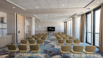 Uma sala de conferências com fileiras de cadeiras, uma tela grande e piso coberto de carpete.