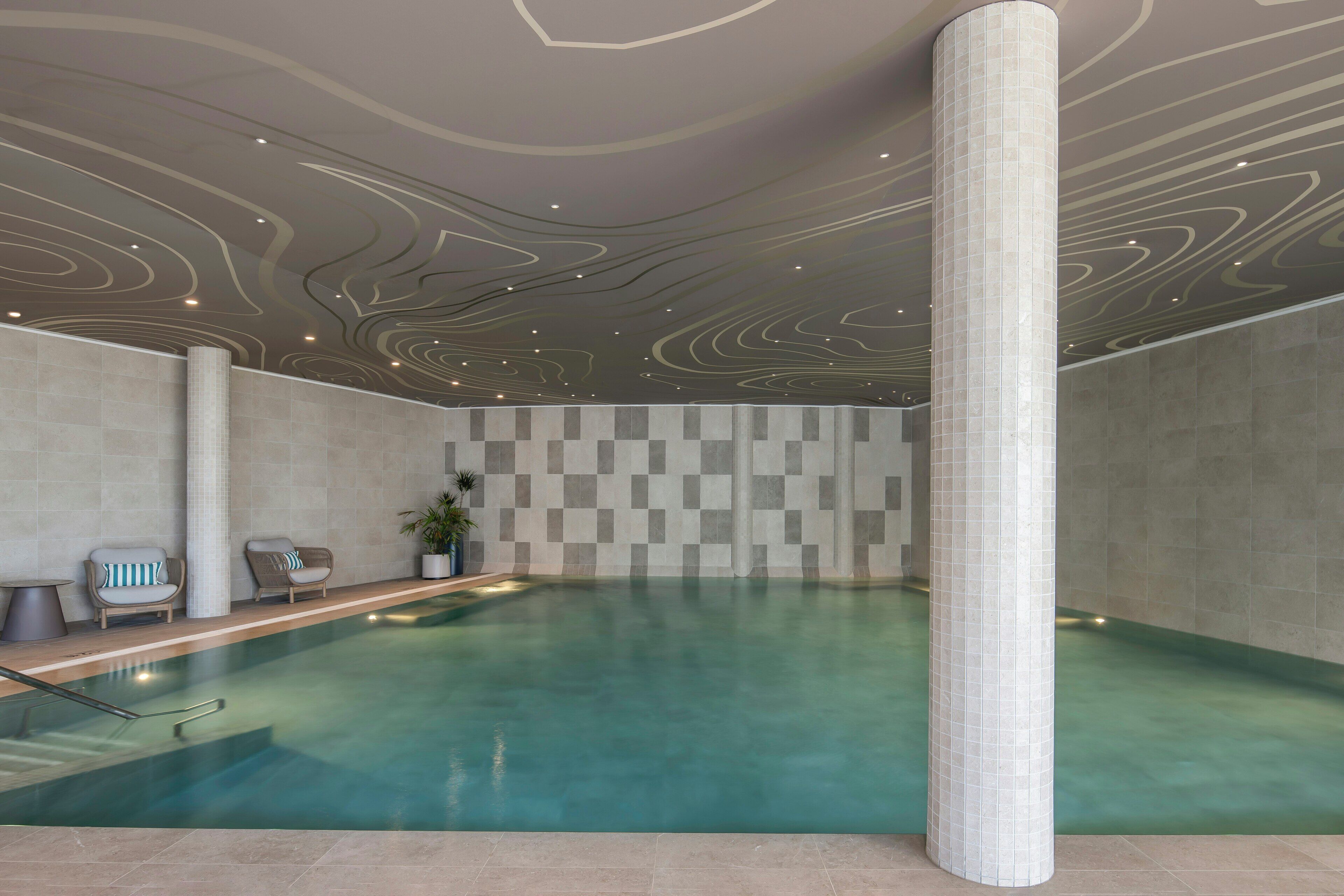 Une piscine intérieure avec un mur orné d’un motif à carreaux, un espace détente avec des fauteuils et un plafond décoratif.