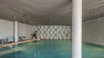 Une piscine intérieure avec un mur orné d’un motif à carreaux, un espace détente avec des fauteuils et un plafond décoratif.
