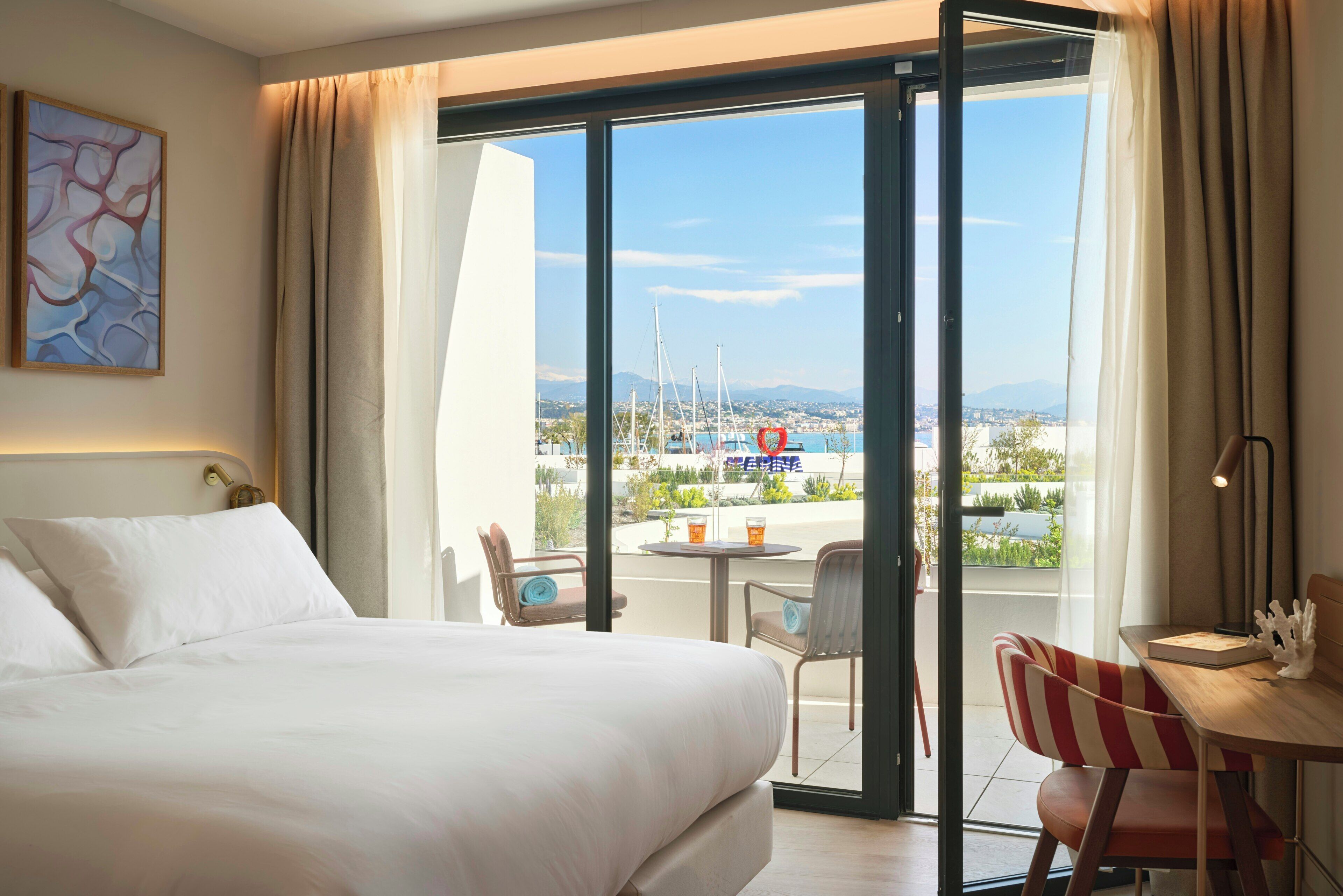 Une chambre d’hôtel avec un lit, une chaise, une table et une vue sur la mer et les bateaux.