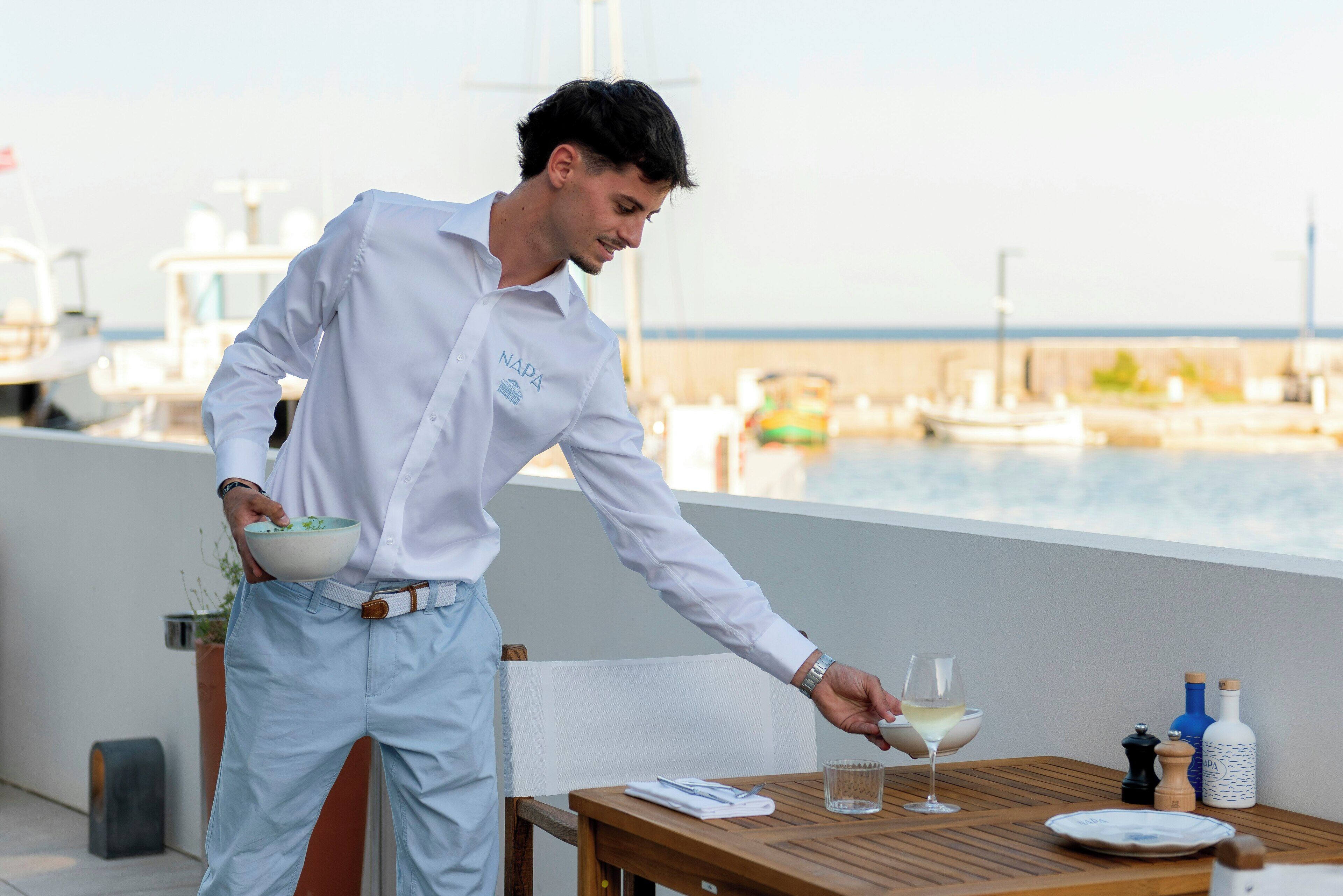 Un hombre sirve una bebida sobre una mesa de madera, con vista al puerto deportivo de fondo.