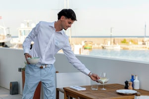 Un hombre sirve una bebida sobre una mesa de madera, con vista al puerto deportivo de fondo.