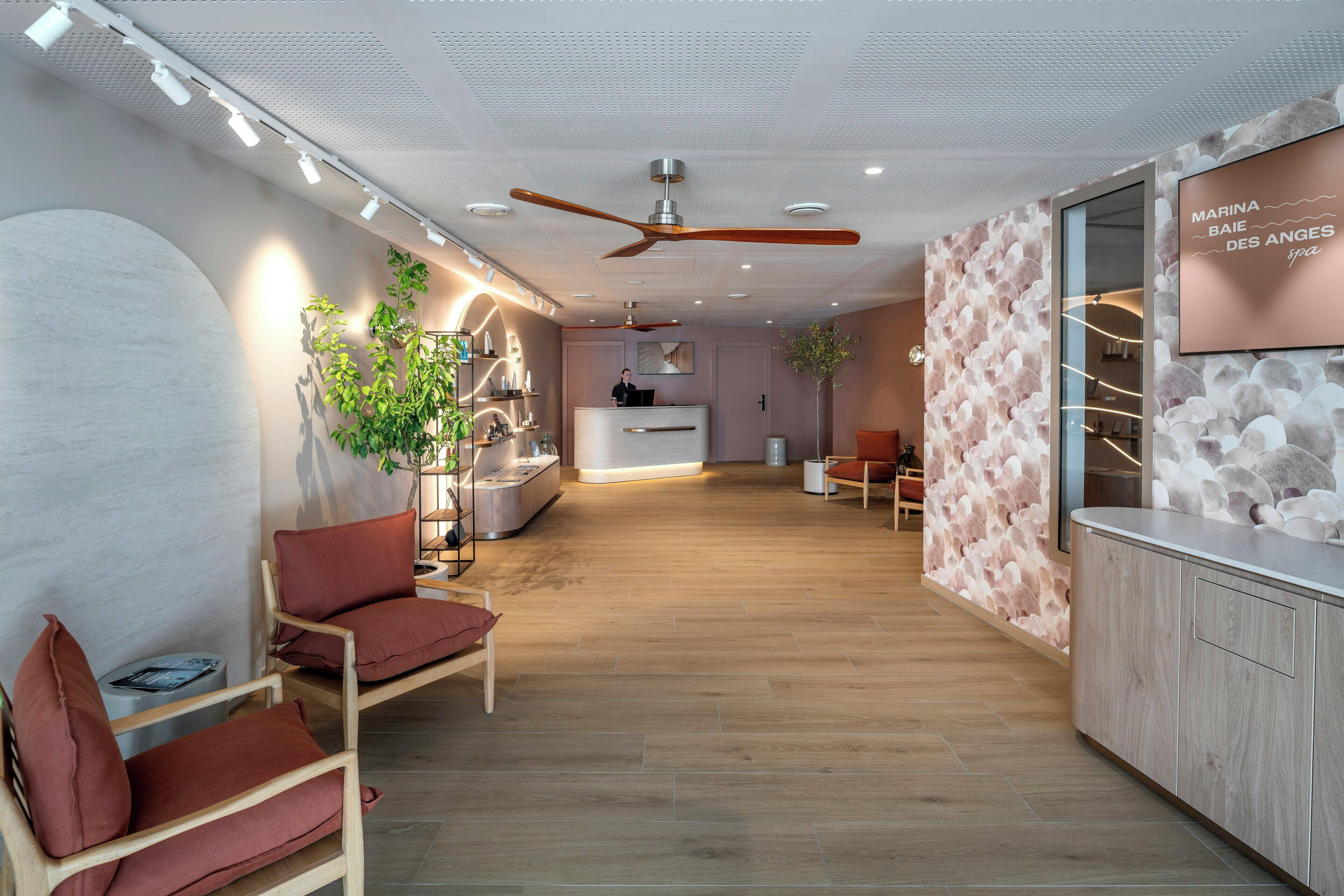 Zona de recepción moderna con un mostrador de madera, área de estar con cojines rojos y un televisor empotrado mostrando el nombre del spa.