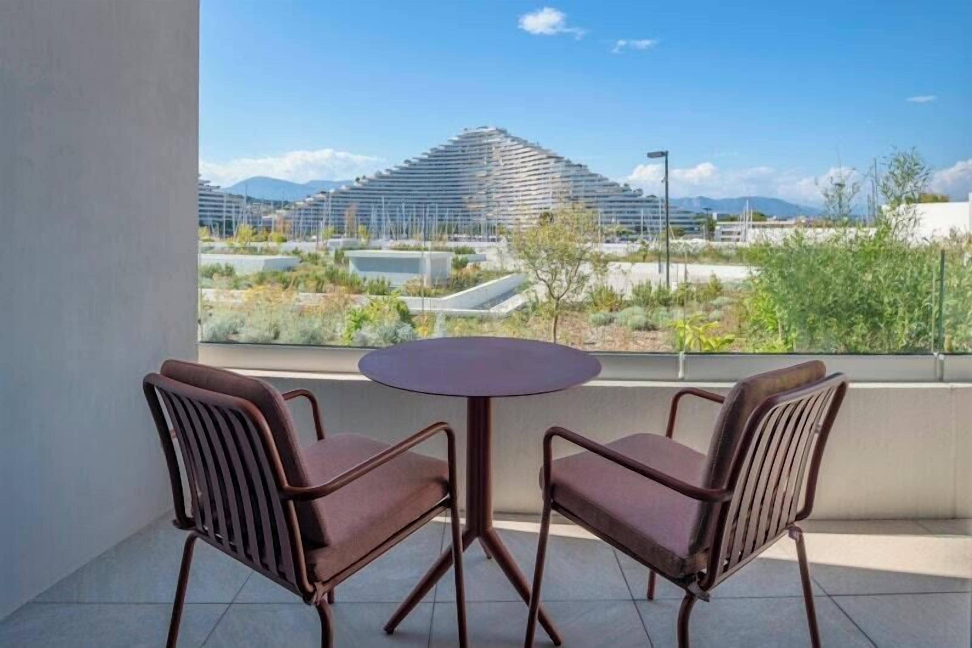 Un balcon avec deux chaises et une table, donnant sur un bâtiment moderne et un espace paysager aménagé.