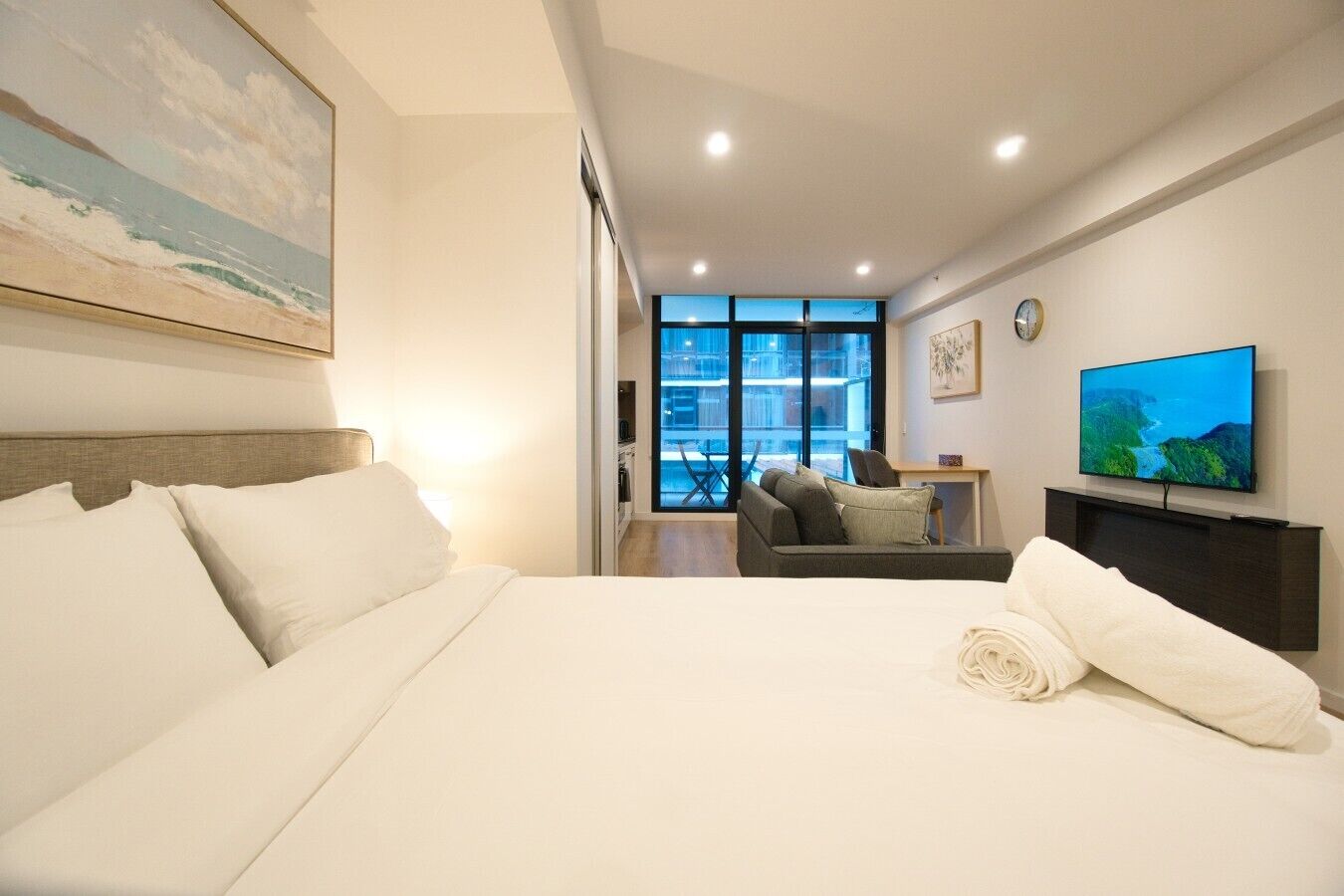 A hotel room with a large bed, a television, a desk, and a view of the city through large windows.