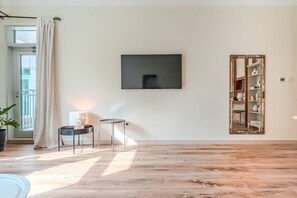Ein Zimmer mit Holzboden, einem schwarzen Spiegelrahmen, einem kleinen Tisch mit Lampe und einer Tür mit weißem Vorhang.