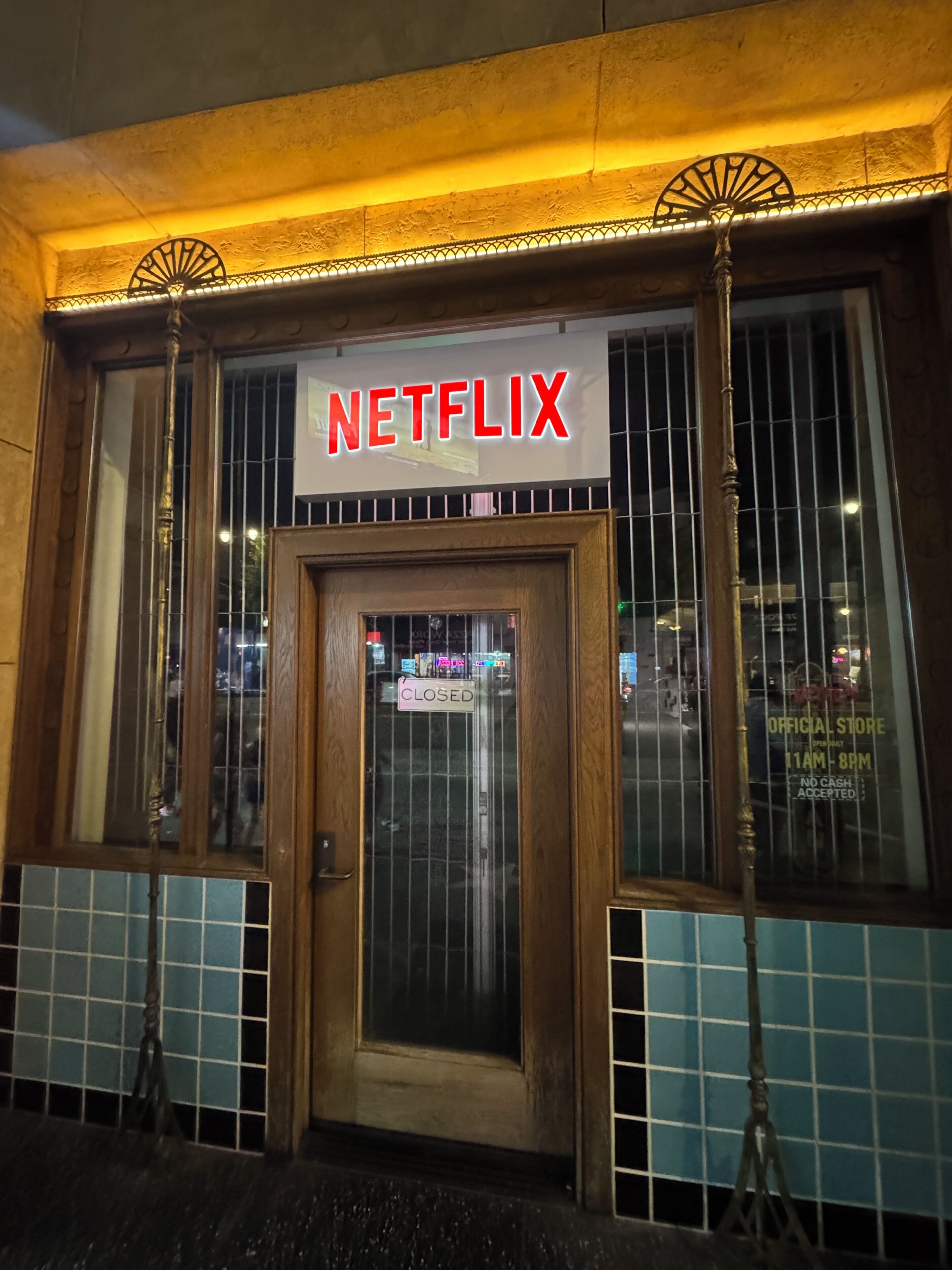 Een gebouw met een bordje met de tekst 'NETFLIX' boven de ingang, een gesloten bord op de deur en een raam met een rasterpatroon.
