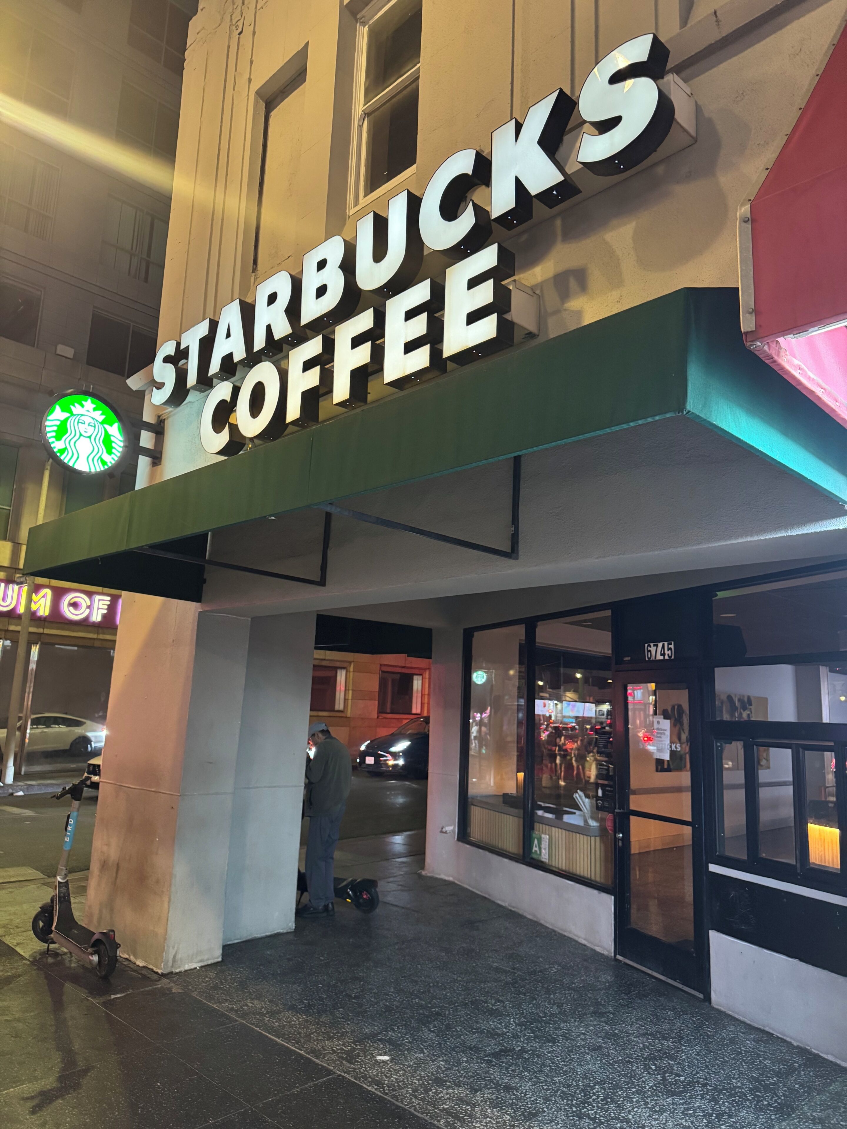 Een Starbucks Coffee winkel met een groot verlicht bord, een duidelijke ingang en een scooter die er buiten geparkeerd staat.