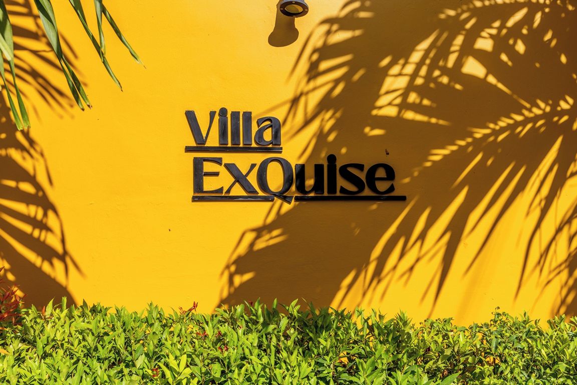Eine gelbe Wand mit dem Schriftzug 'Villa ExQuise' und dem Schatten eines Palmenbaums.