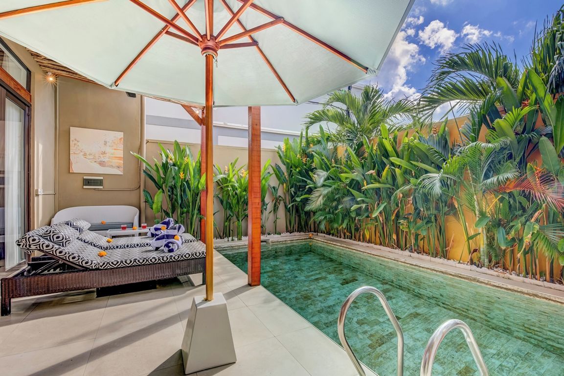 Zona de piscina con una silla de descanso, una sombrilla grande y exuberante vegetación tropical.