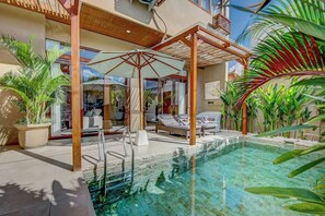 A pool area with a sun umbrella, lounge chairs, and a view of the interior. - Villa Exquise (Legian)