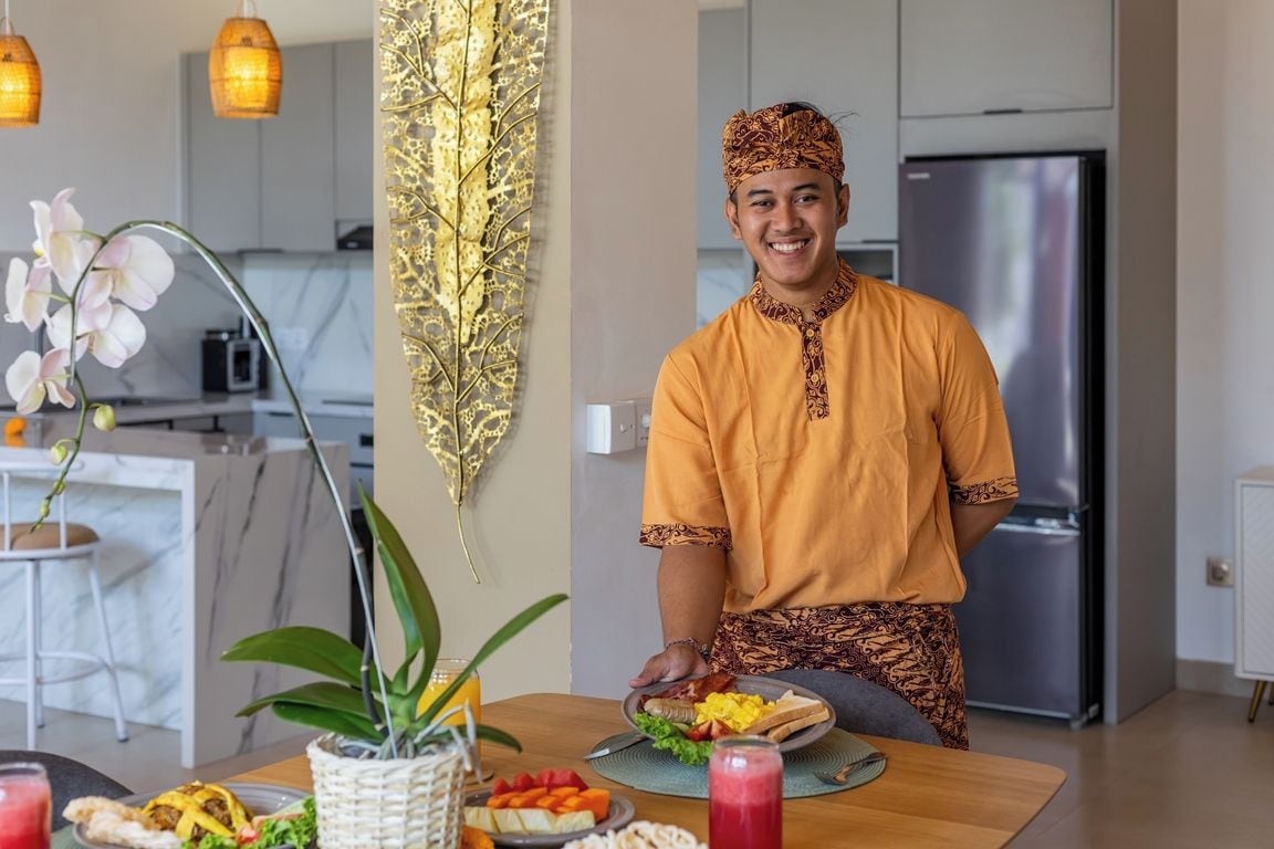 Una persona vestida con traje tradicional sirve comida en una cocina moderna con refrigerador y luces colgantes.