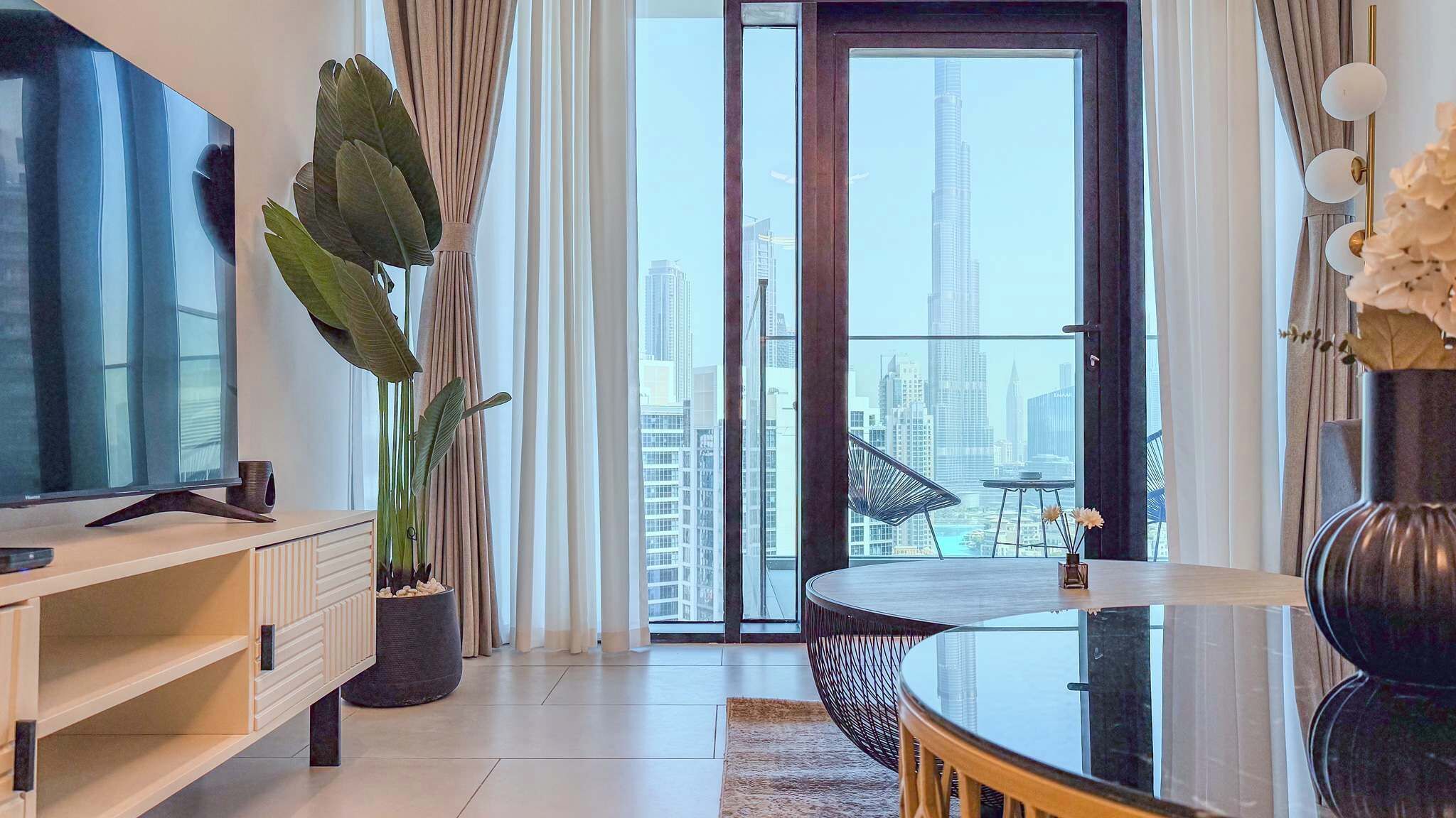 A modern living room with a flat-screen TV, a potted plant, a round glass table, and a view of a cityscape through a large window.