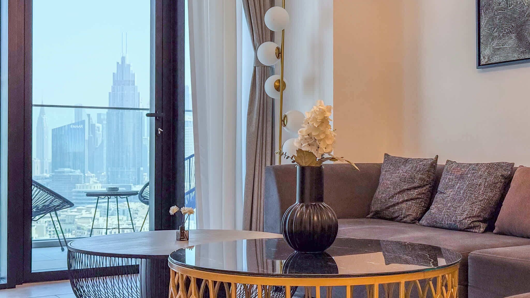 Una sala de estar moderna con una mesa de centro redonda de vidrio, un jarrón oscuro con flores, un sofá gris con cojines estampados y vistas a un horizonte urbano a través de amplias ventanas.