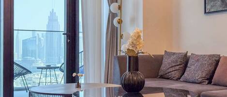 Un salon moderne avec une table basse ronde en verre, un vase foncé orné de fleurs, un canapé gris avec des coussins à motifs et une vue sur les toits d’une ville, visible à travers de grandes fenêtres.