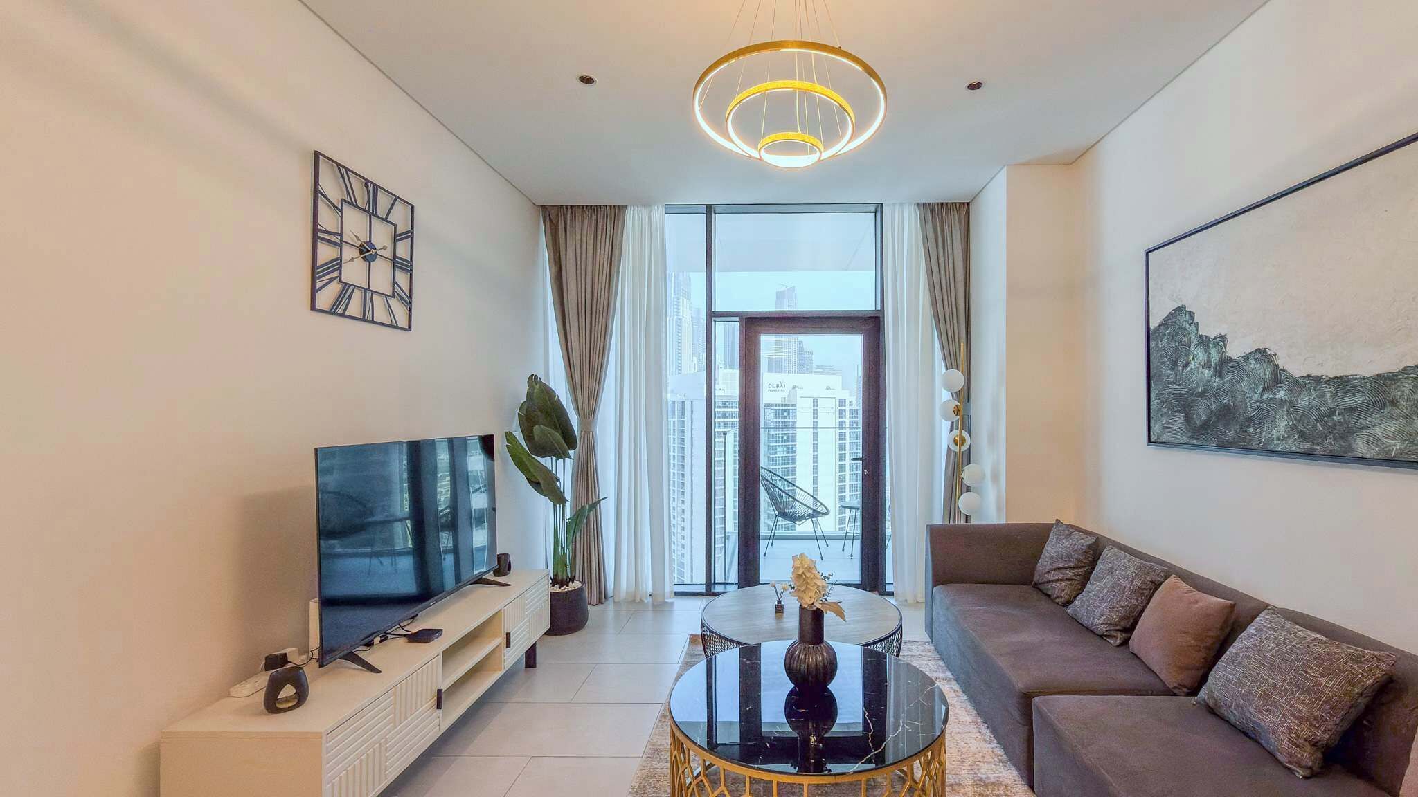 Un salon moderne doté d’un téléviseur à écran plat, d’une table basse ronde, d’un canapé et d’une grande fenêtre offrant une vue sur la ville.