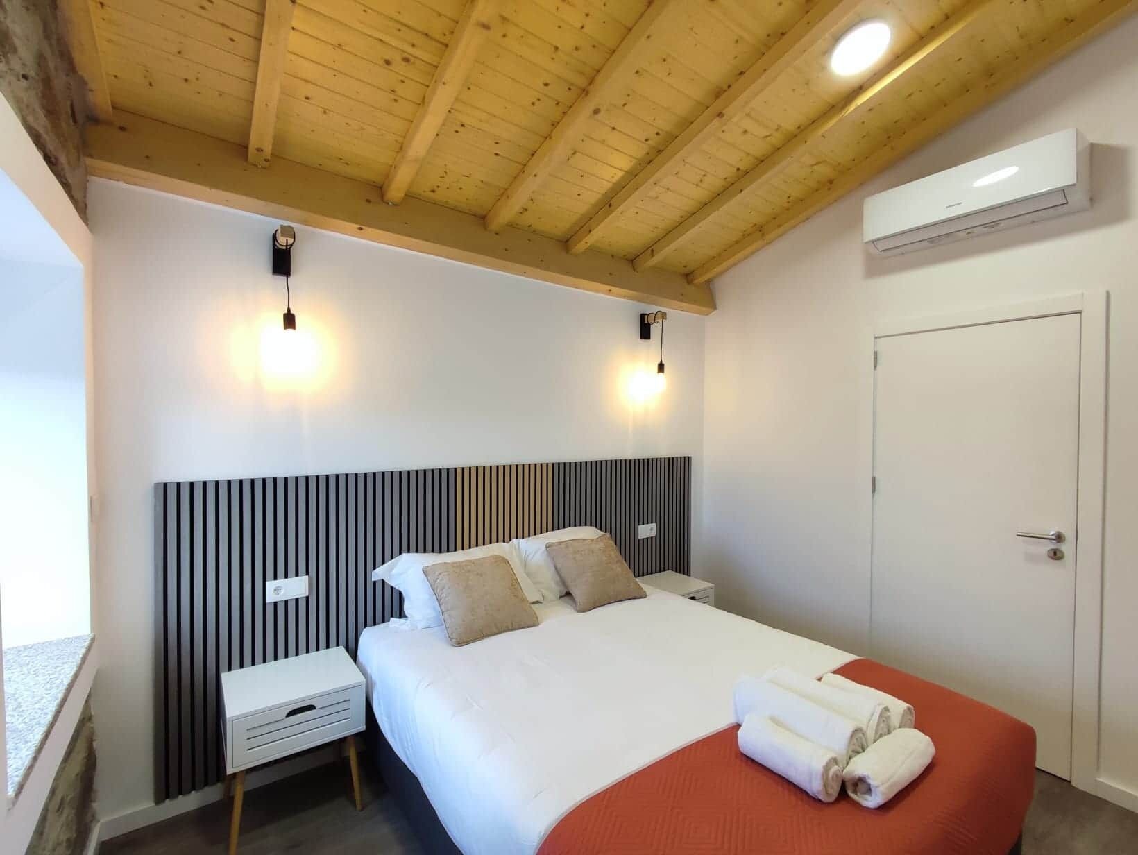 A hotel room with a bed, bedside tables, a wooden ceiling, and an air conditioning unit.