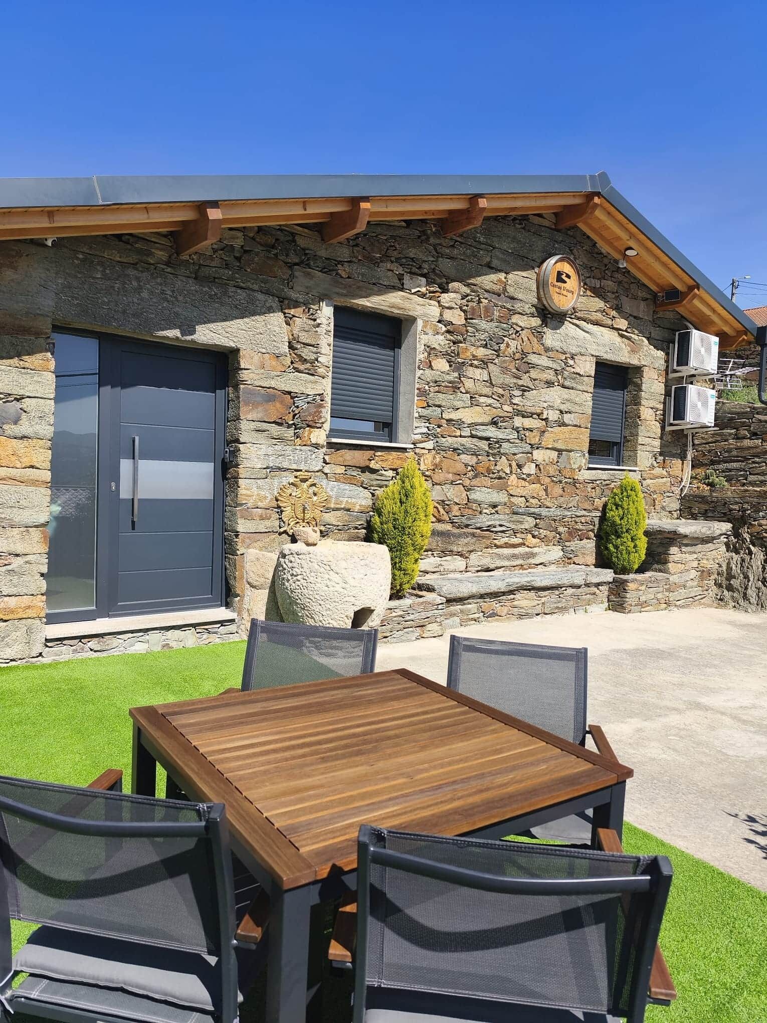 A stone building with a wooden roof, a door, and a window. There is a table and chairs in front of the building.
