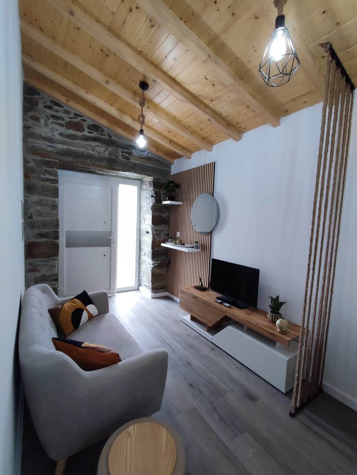 A modern living room with a stone wall, wooden ceiling, a grey sofa, a wooden TV stand, and a hanging light fixture.