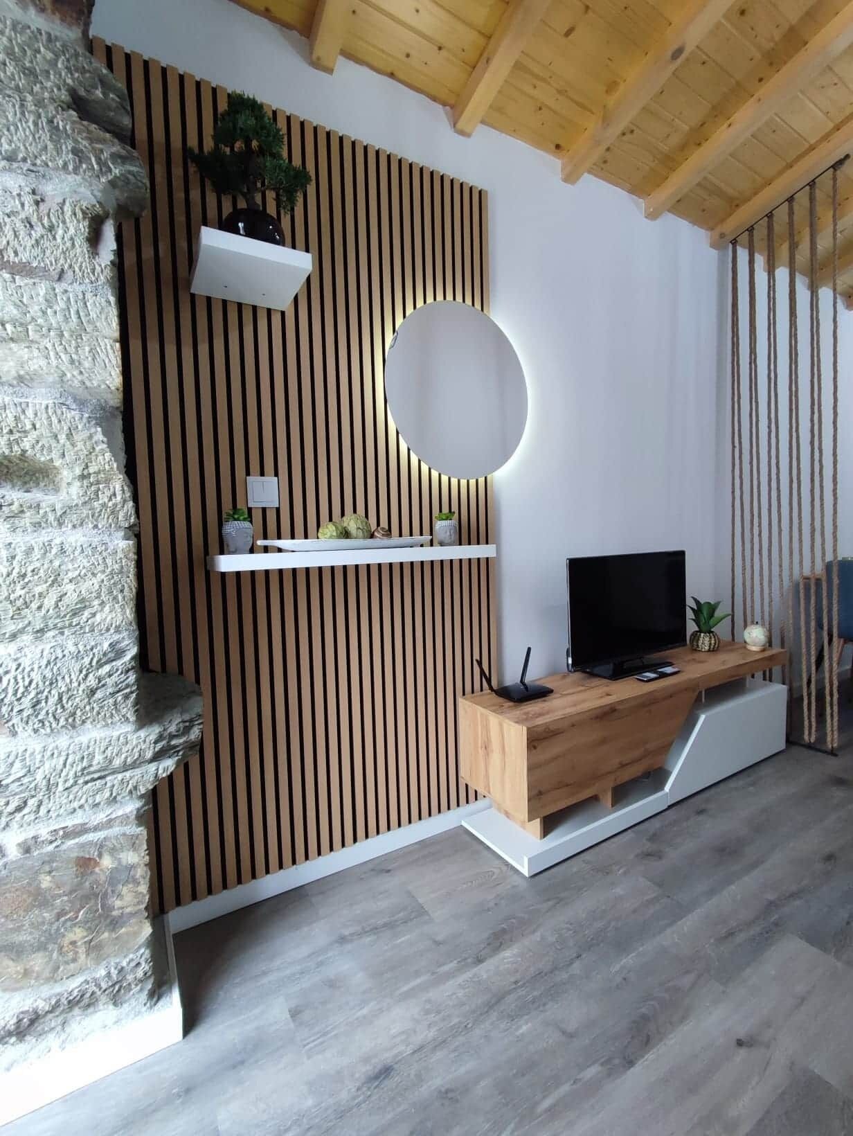Una sala moderna con pared de piedra decorativa, listones de madera, un espejo redondo, un televisor de pantalla plana y un mueble bajo de madera.