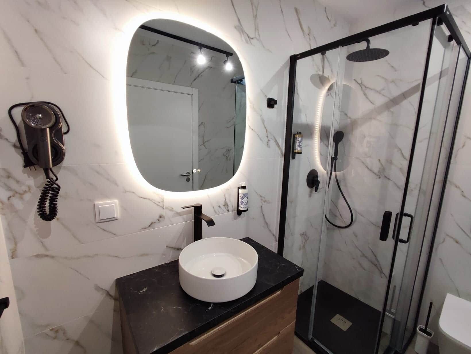 A bathroom with a marble wall, a round sink, a mirror, a hairdryer, and a glass-enclosed shower.