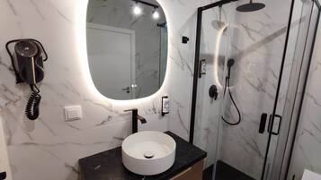 A bathroom with a marble wall, a round sink, a mirror, a hairdryer, and a glass-enclosed shower.