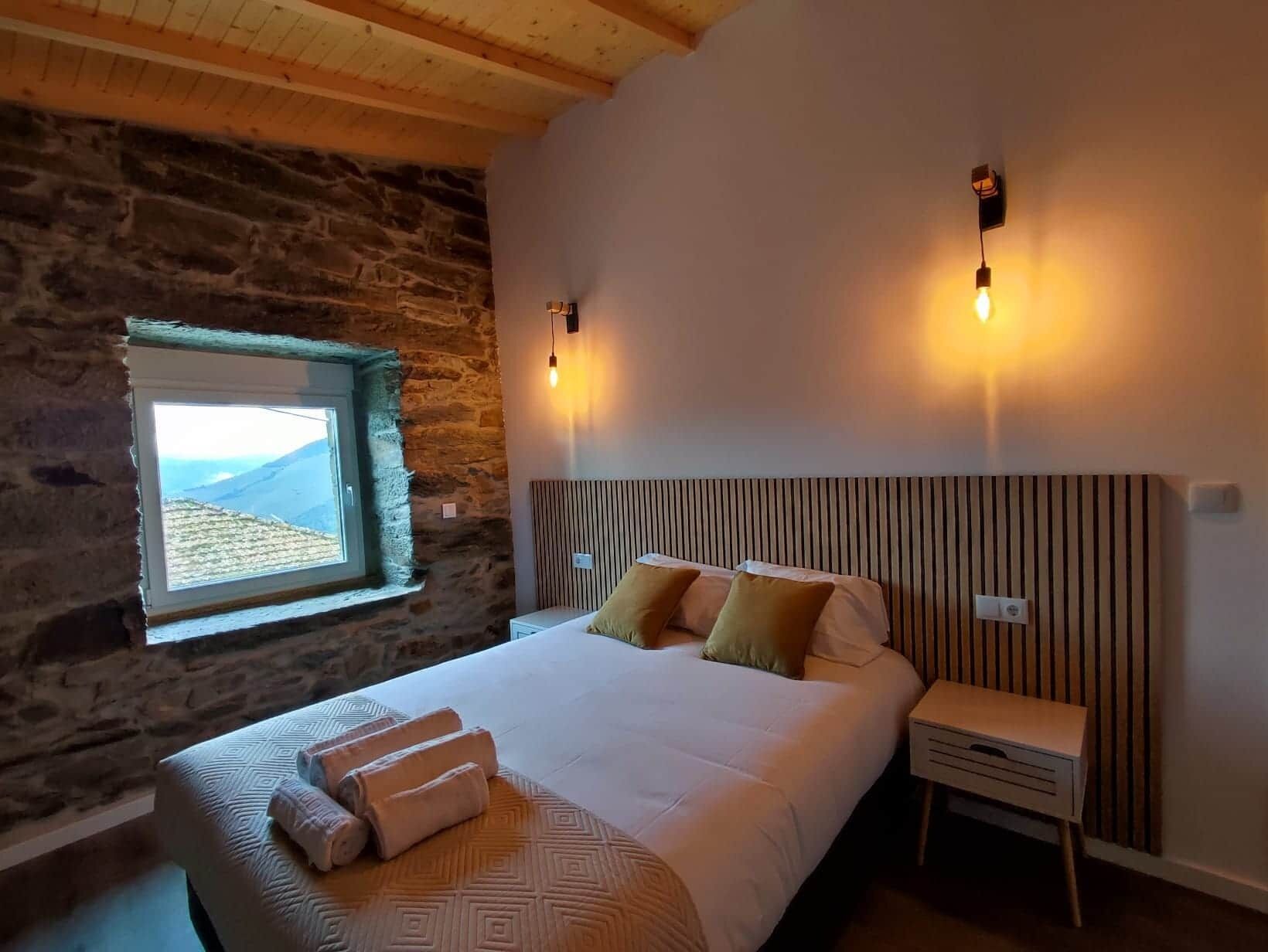 A bedroom with a stone wall, a window with a view, a bed with pillows, and bedside lamps.