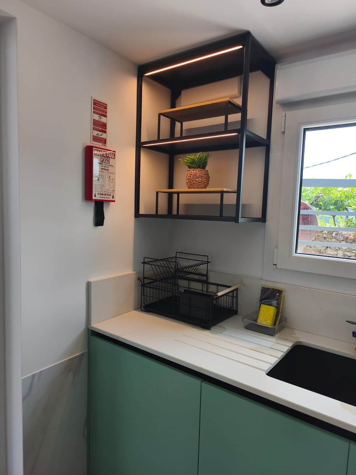 A kitchen with a white countertop, green cabinets, a black metal shelf unit, and a window with a view of greenery.