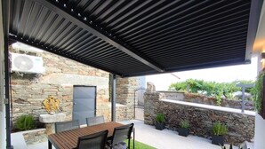 A covered patio with a stone wall, a wooden table with chairs, and potted plants. - Castas D'ouro Guest House II (Casal de Loivos)