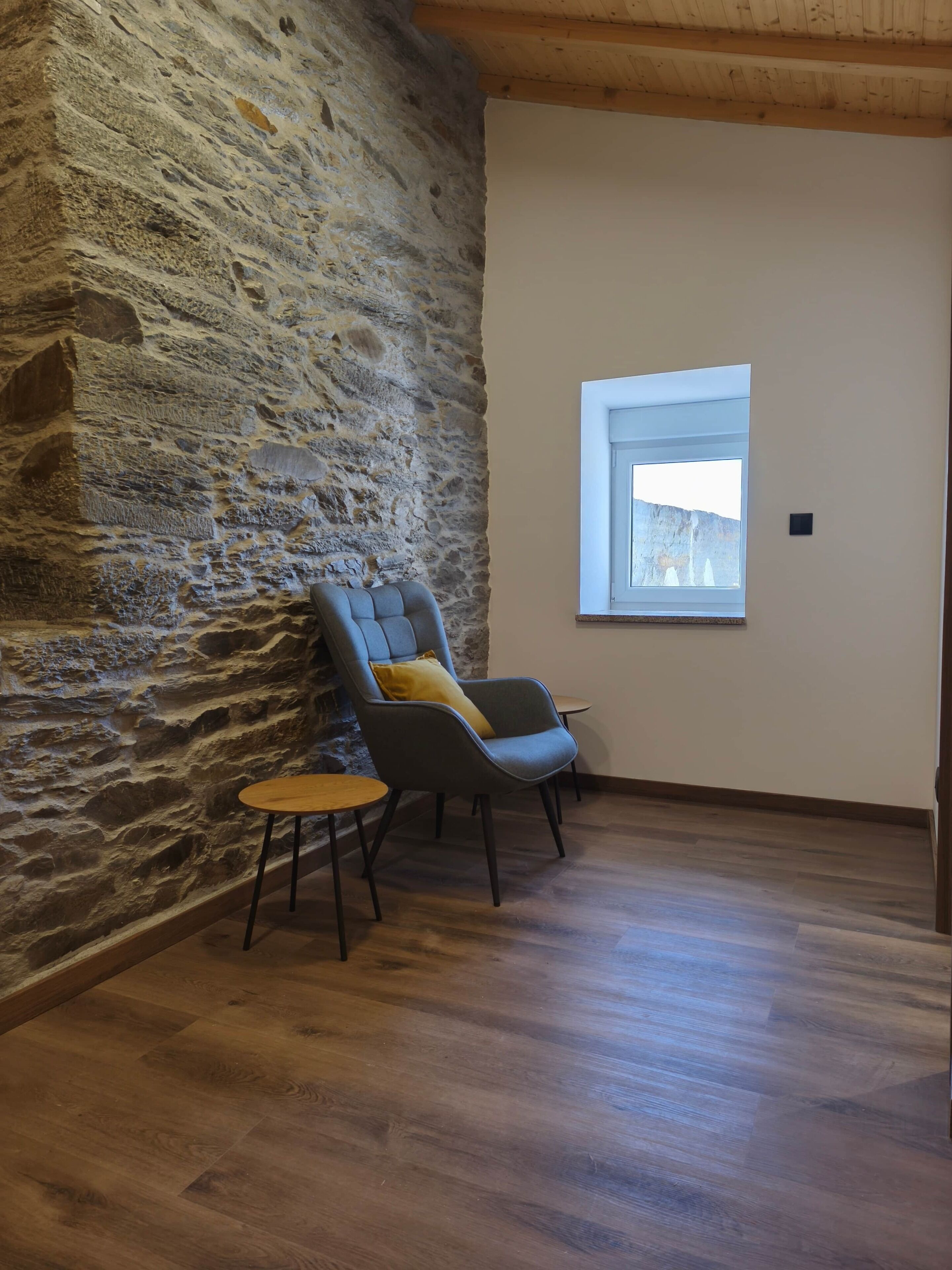 A room with a stone wall, a wooden floor, a window, and a chair with a small table.
